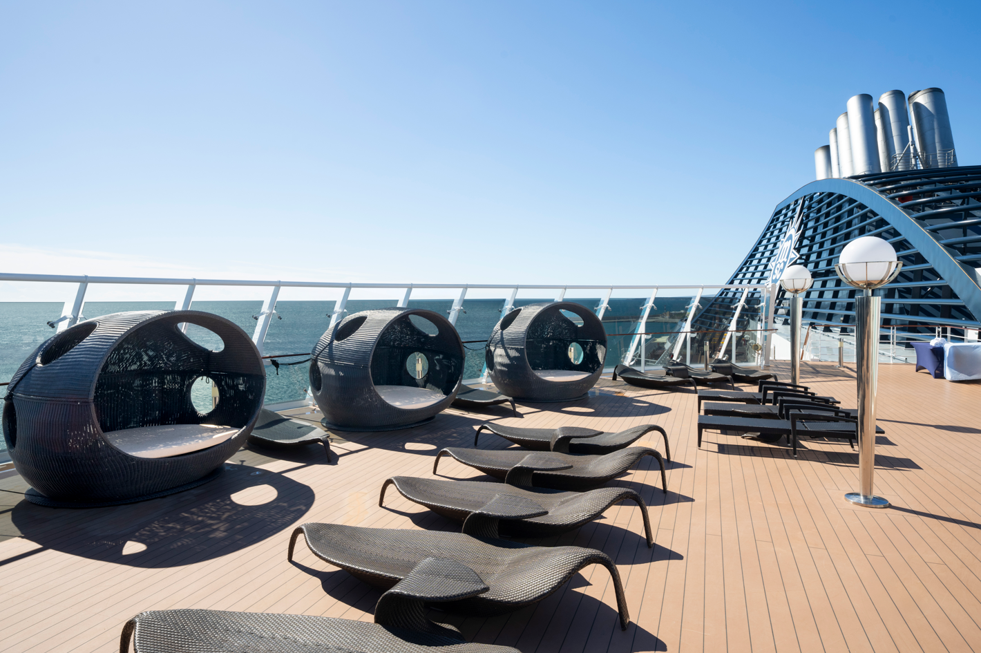 The image shows the deck of a cruise ship featuring a series of modern lounge chairs and circular seating arrangements. The scene is bright and sunny, with a view of the ocean in the background.