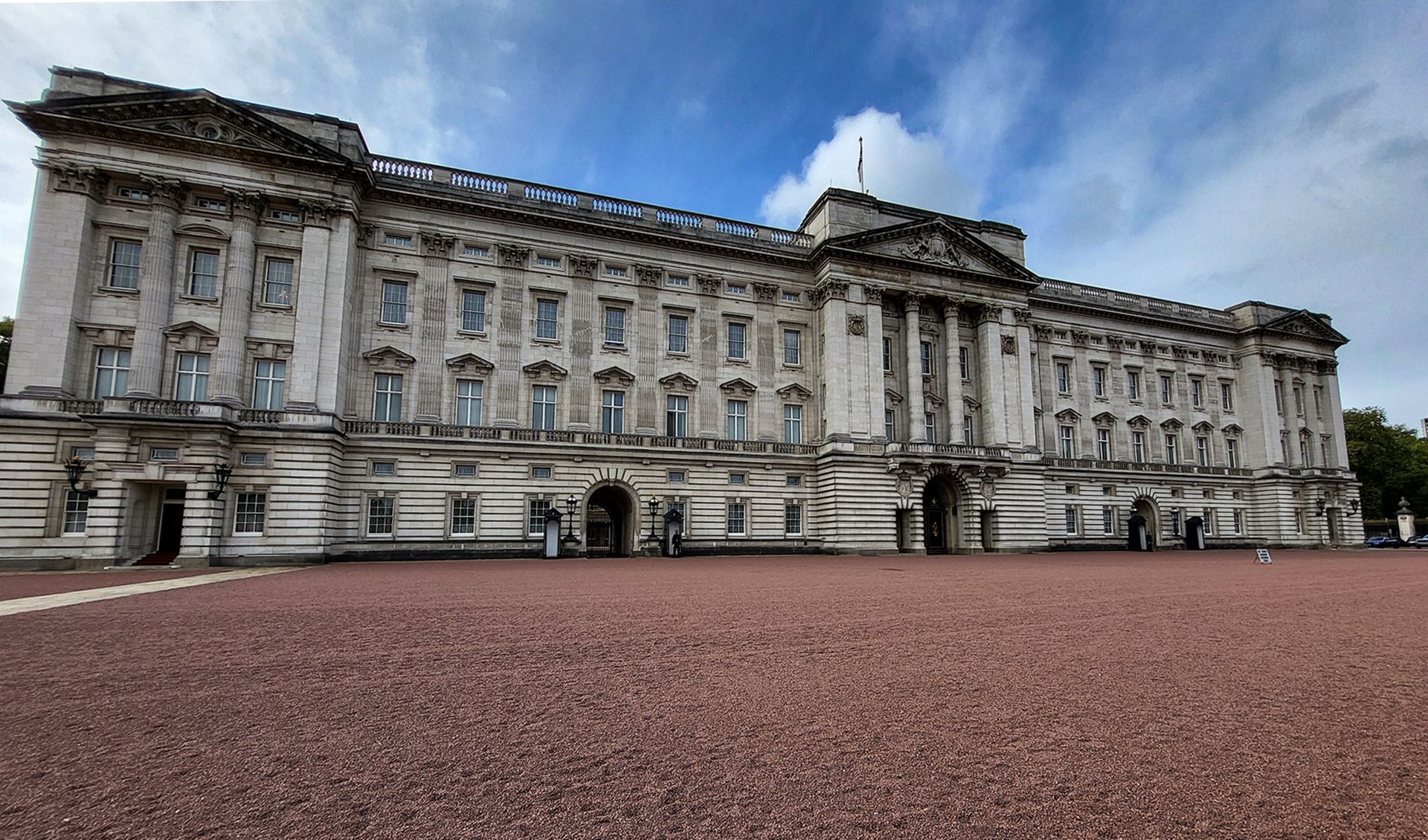 An exterior view of Buckingham Palace in London, showcasing its grand architecture and the surrounding courtyard. The sky appears to be partly cloudy, adding to the atmospheric setting.