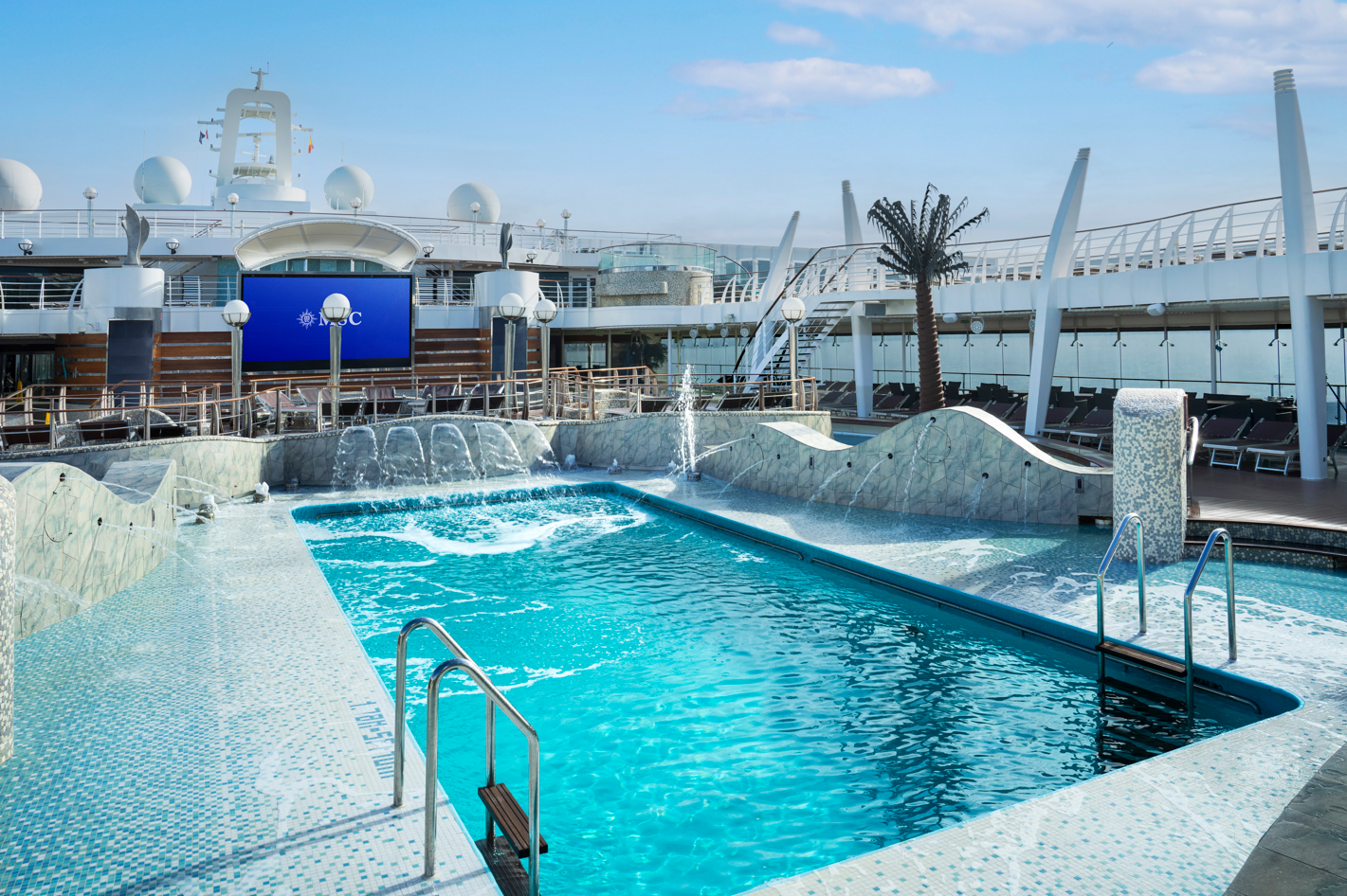 The image shows a luxurious swimming pool area on a cruise ship, featuring modern design elements and clear blue water. Sun loungers are arranged around the pool, with a scenic view of the ocean in the background.