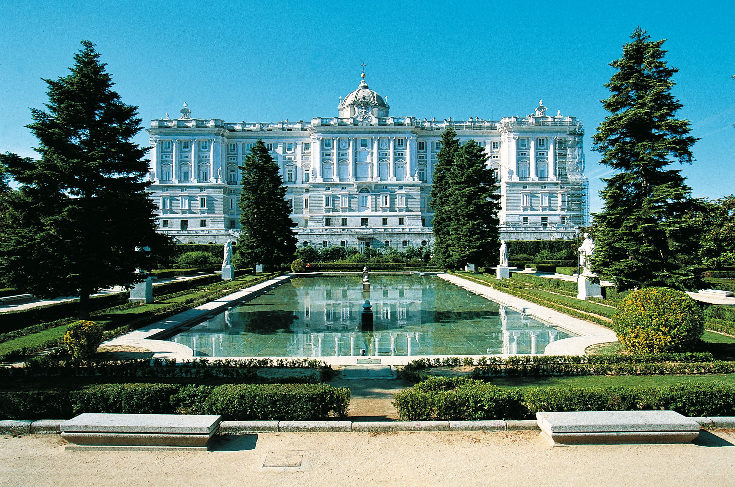 The image showcases the Royal Palace of Madrid, an architectural marvel surrounded by lush greenery and a serene reflecting pool. The palace features ornate facades and a prominent dome, set against a clear blue sky.