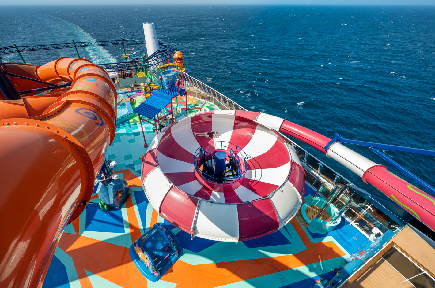 The image showcases the deck of a cruise ship featuring colorful water slides and a lively water park area. The vibrant design and ocean backdrop create an inviting atmosphere for leisure activities.