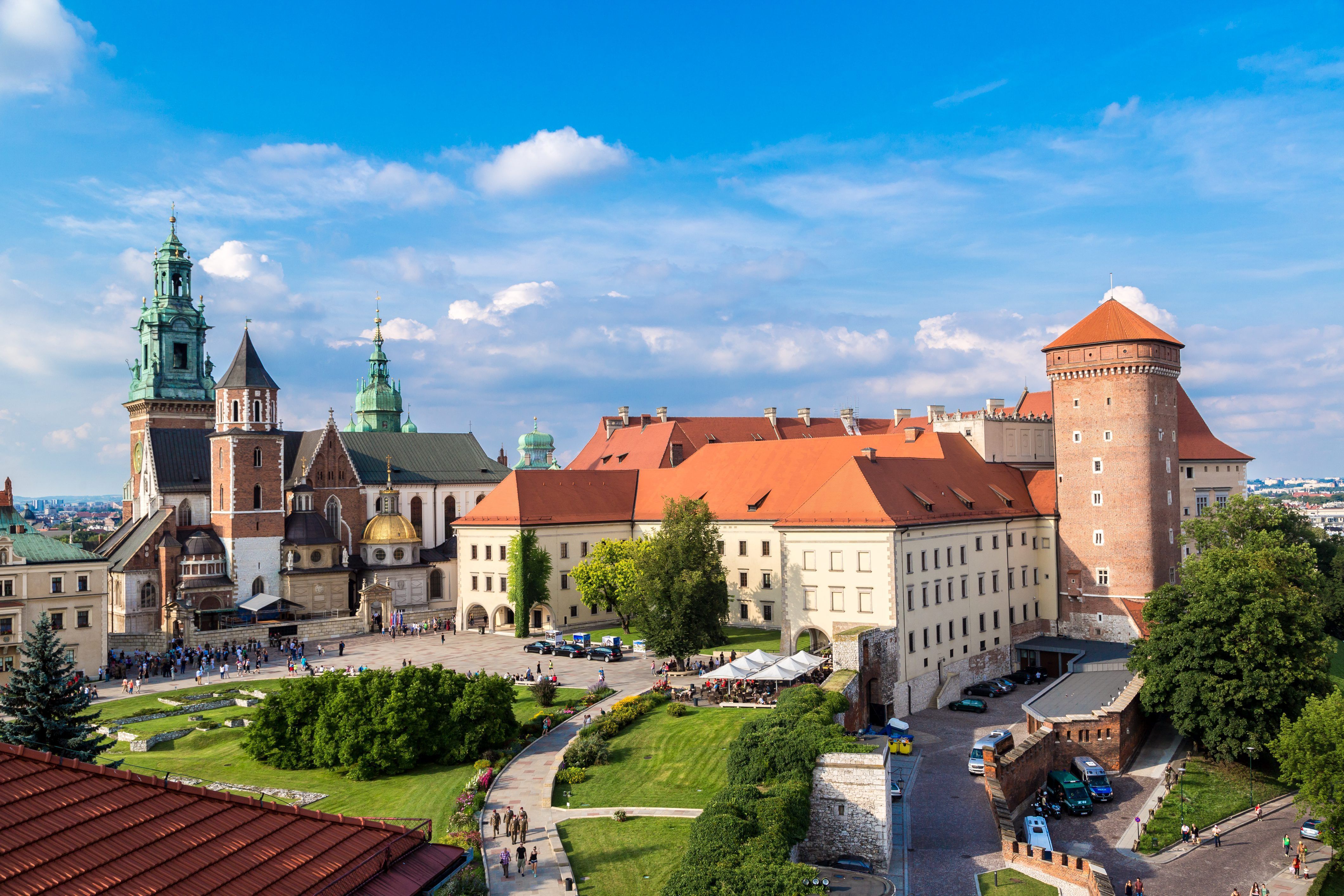 The image showcases Wawel Castle, a historic site located in Kraków, Poland. It features a panoramic view of the castle complex, highlighting its distinctive architectural styles and lush surrounding greenery under a clear blue sky.