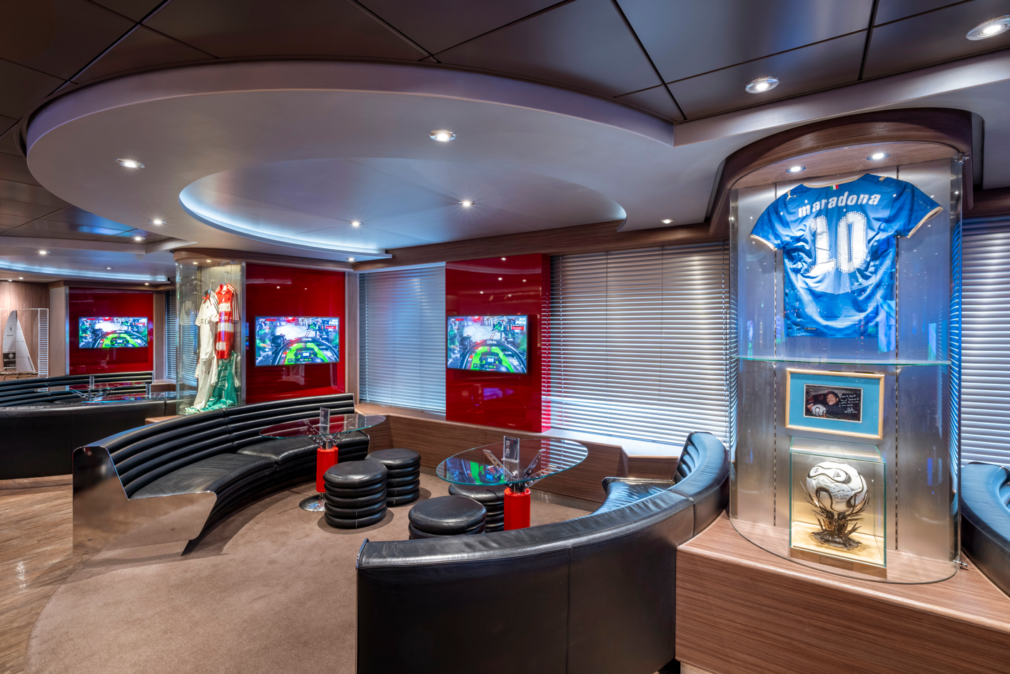The image depicts a stylish interior of a lounge or private room featuring a display case with sports memorabilia, including a jersey hung prominently. Two televisions are visible, with a vibrant red backdrop enhancing the modern aesthetic.