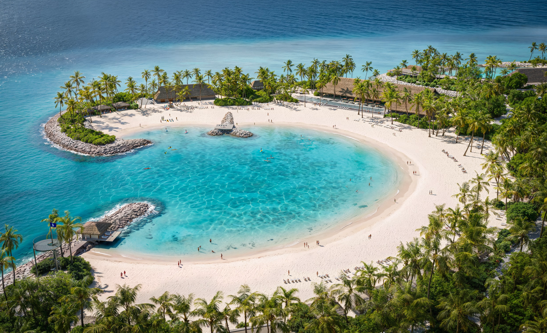Aerial view of a tropical beach lagoon featuring clear turquoise waters, surrounded by lush palm trees and a sandy shoreline. A few people can be seen enjoying the beach, with palm-fringed huts in the background.